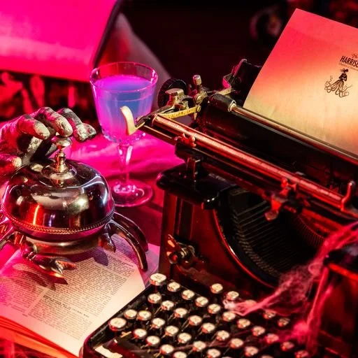 A plethora of vintage objects, including a typewriter, reception bell, and blue cocktail, all on a red-lit surface.