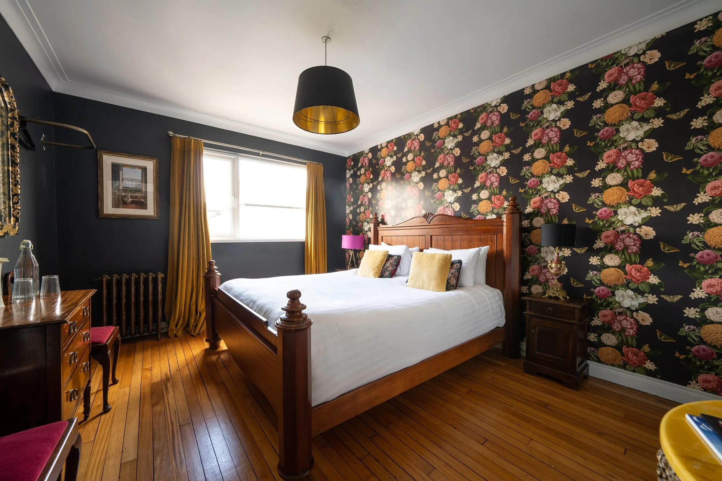 A bedroom with a wooden bed, floral accent wall, yellow curtains, black pendant light, and wooden flooring.