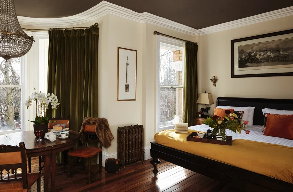 A bedroom with a dark wooden bed, white and orange bedding, and a tray of flowers on the bed. There is a wooden nightstand with a lamp and books, and large windows with green curtains allowing natural light.
