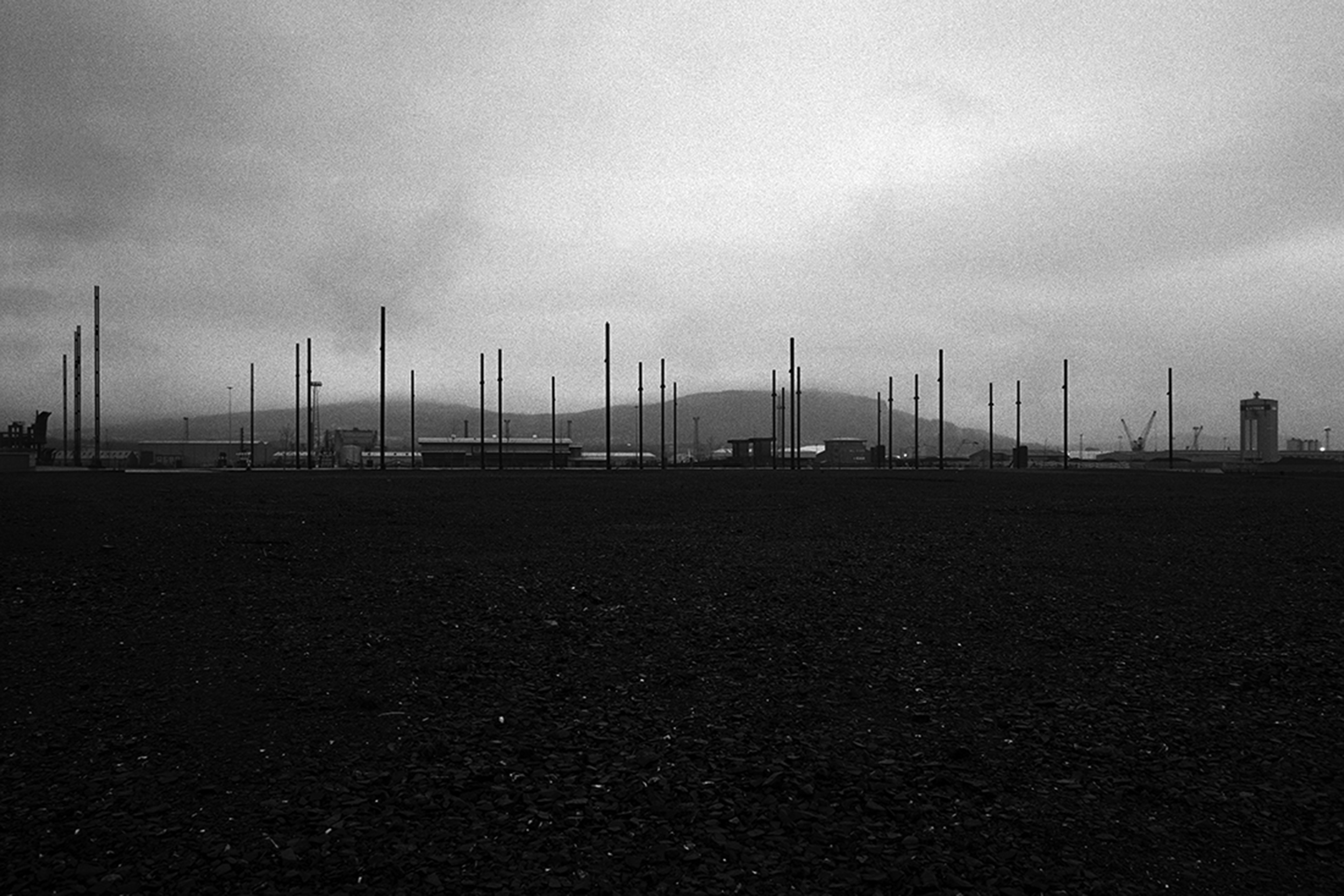 An empty outdoor area with scattered tall poles, industrial buildings, and a mountain in the background under a cloudy sky.