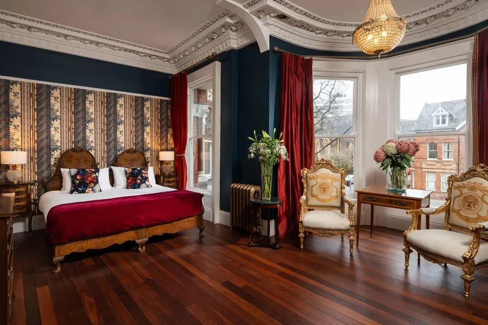 Elegant bedroom with dark blue walls, ornate white ceiling, hardwood floor, and vintage furniture. Features a bed with a floral headboard, red bedspread, and decorative pillows, flanked by lamps. There are large windows with red curtains, and a sitti