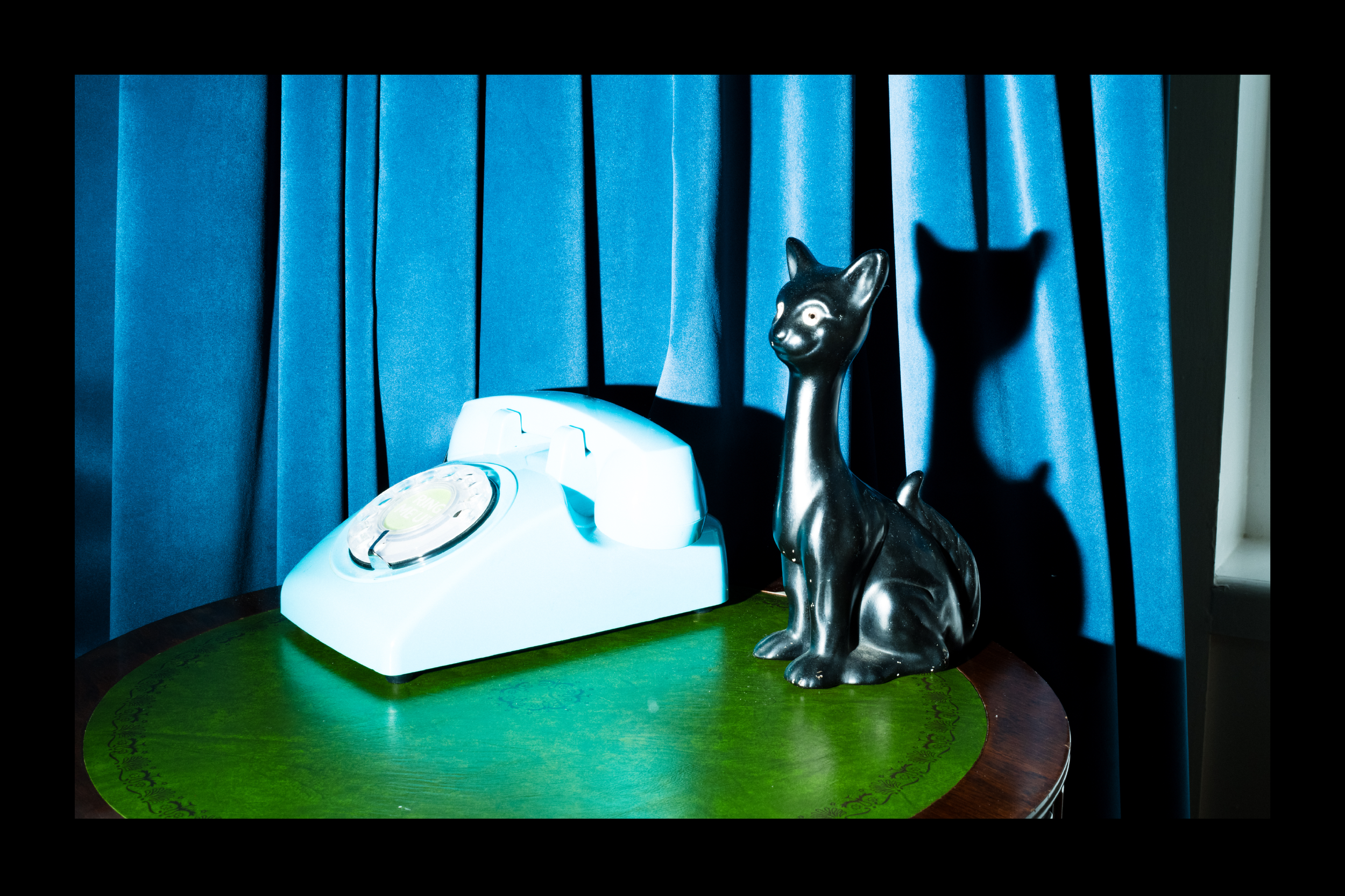 A black cat-shaped sculpture and a white rotary telephone on a green table with blue curtains in the background.