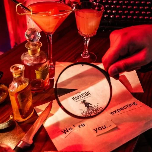 Two cocktails on a bar counter, with a small brown bottle, a knife, and a handwritten note under a magnifying glass. The note reads "HARRISON" and "We're expecting you..."
