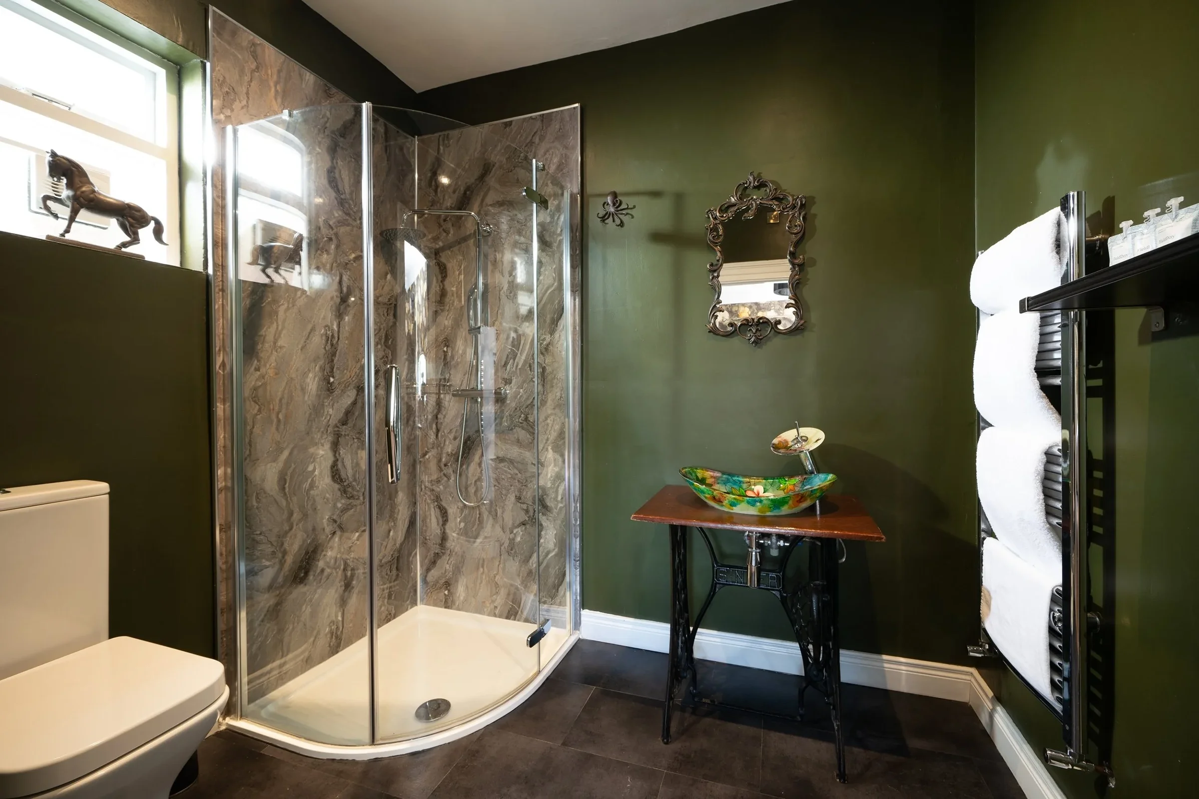 A bathroom featuring a green wall, a glass-enclosed shower with marble walls, a decorative mirror, a vintage-style sink on a wooden table, and a black towel rack with white towels.