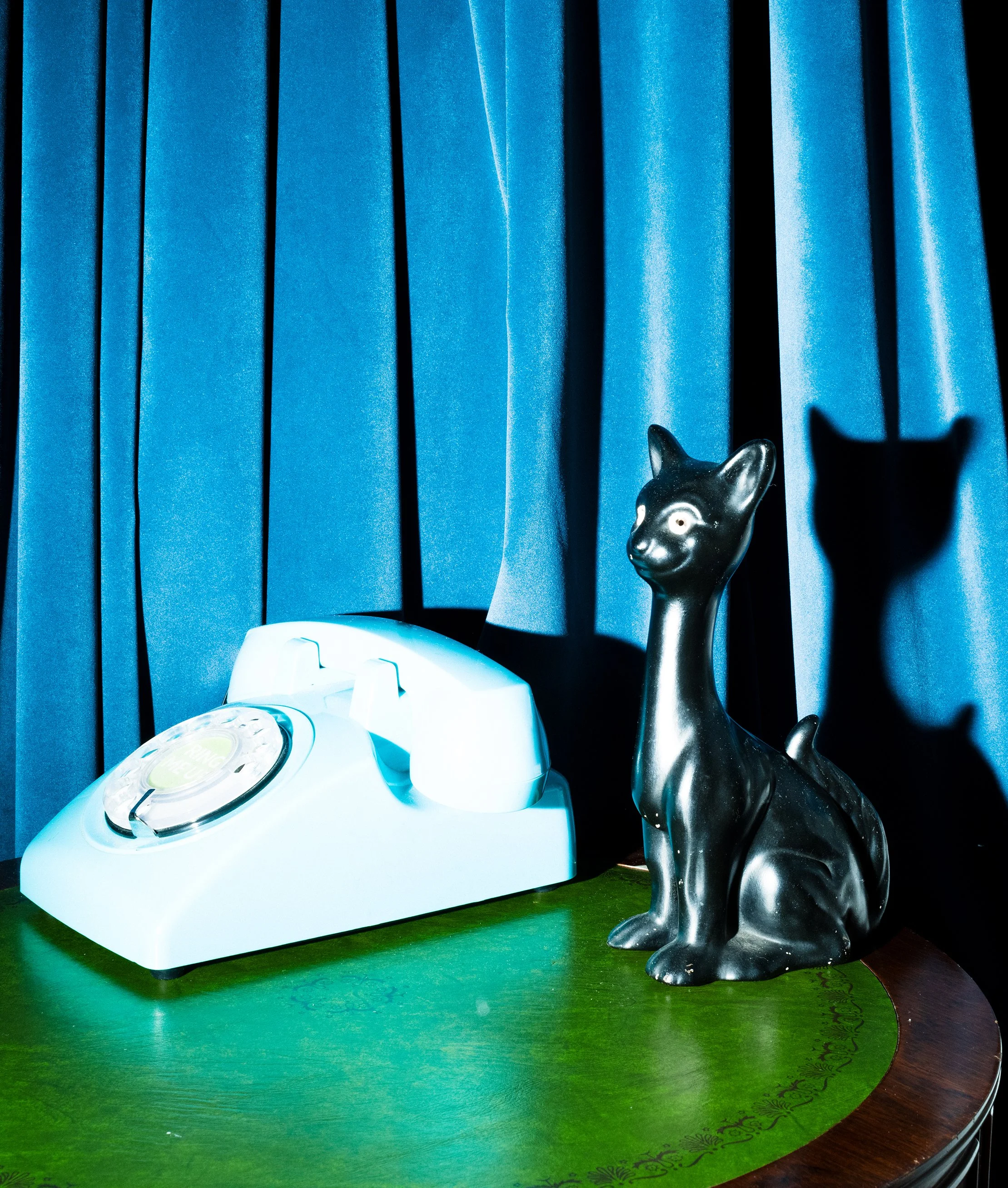 A vintage white rotary telephone and a black cat-shaped figurine on a green wooden table, with blue curtains in the background.