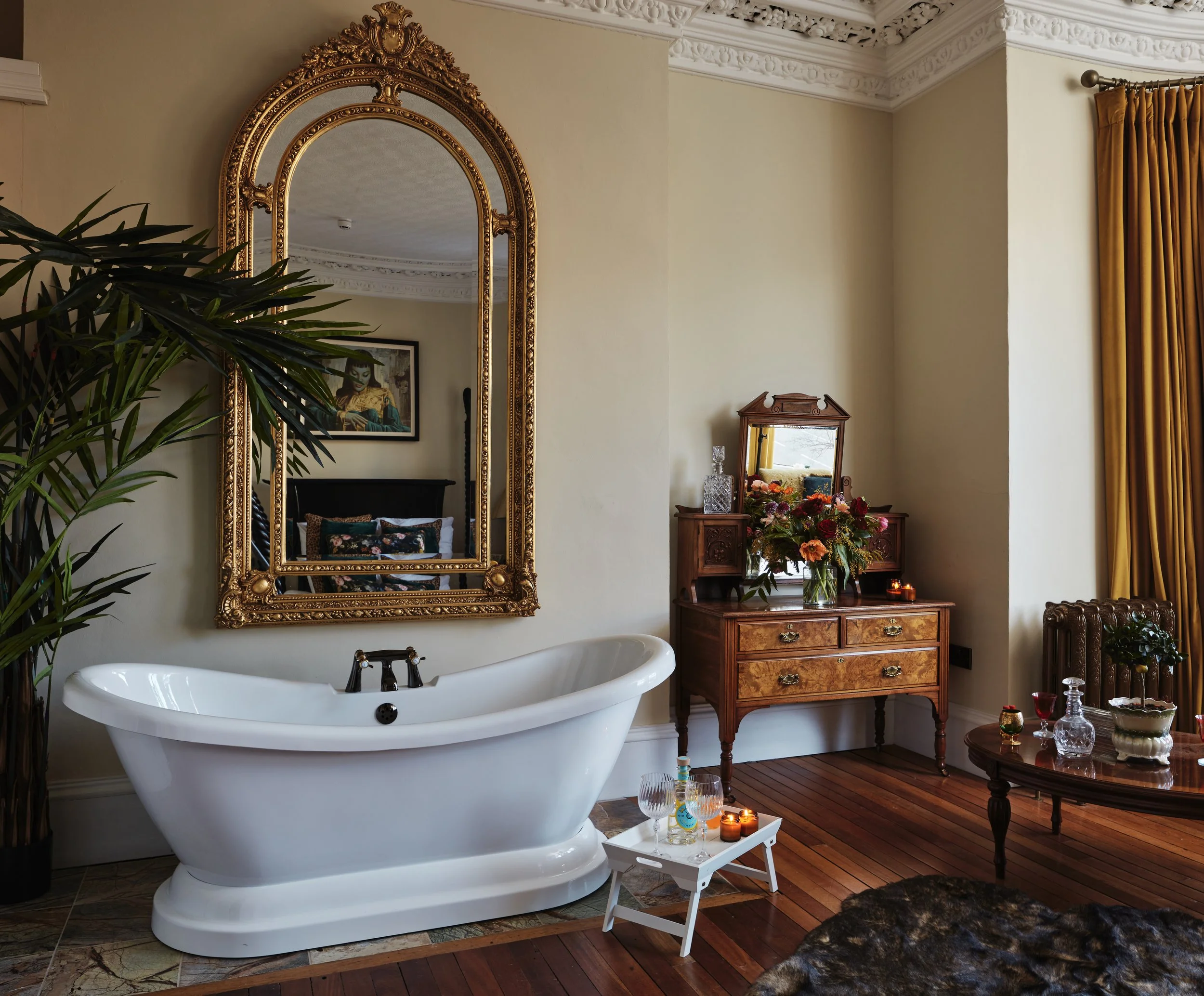 A vintage-style bathroom with a clawfoot bathtub below an ornate gold mirror, a wooden dresser with a mirror holding flower arrangements and decorative glassware, a small tray with glasses and candles, and a round wooden table with glass decanters an