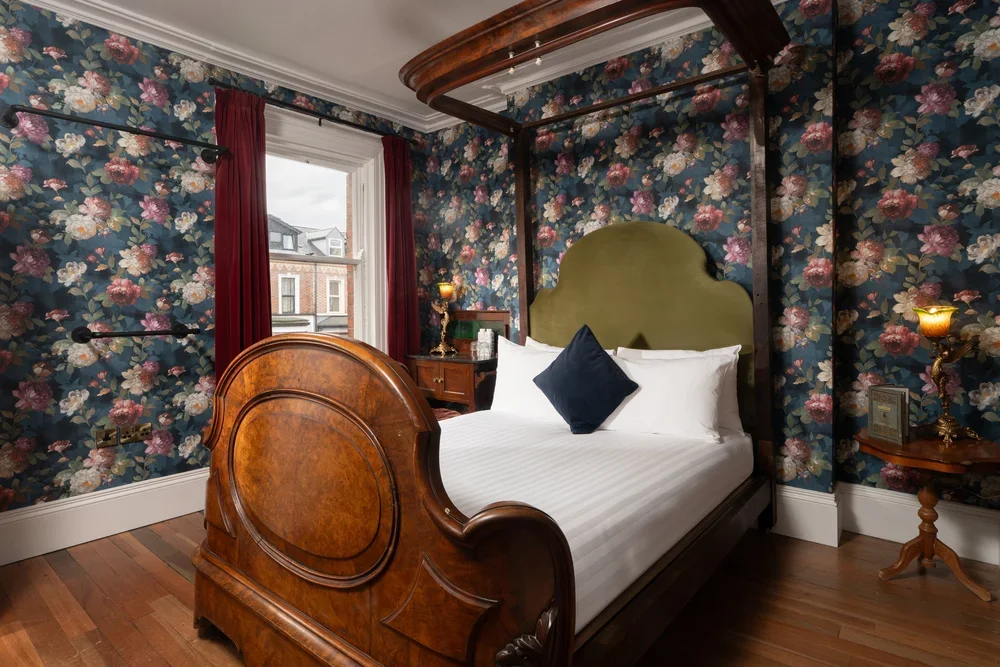 A vintage bedroom with floral wallpaper, a wooden canopy bed with a green headboard, white bedding, a navy pillow, and red curtains. There is a window showing neighboring buildings, a nightstand with a lamp, and hardwood flooring.