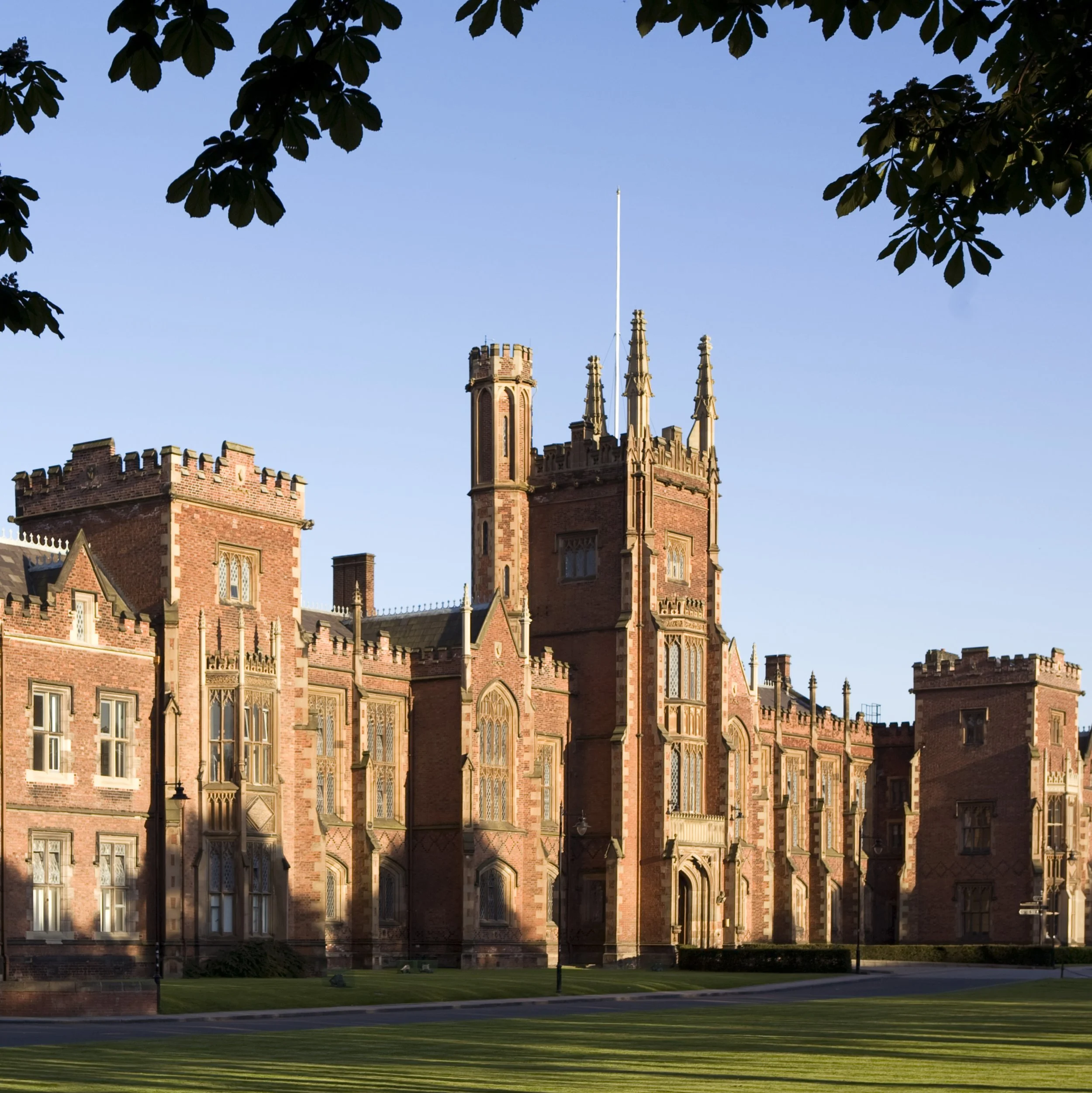 A large, historic red brick castle with towers, crenellated walls, and detailed architectural windows, surrounded by a well-maintained lawn and trees.