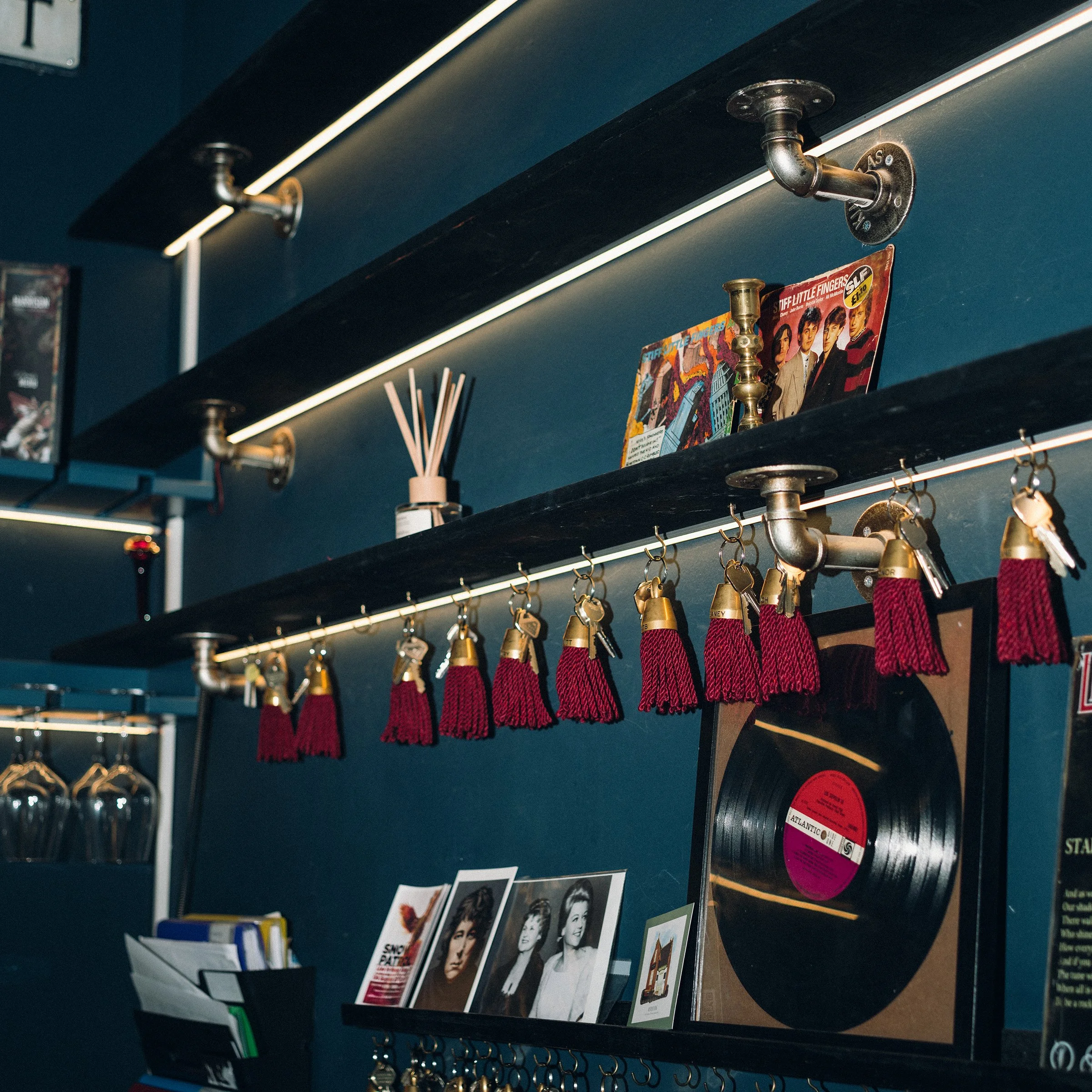 Decorative wall with shelves holding magazines, a candle holder, reed diffuser, framed photos of women, and many red tassel keychains hanging from a pipe.