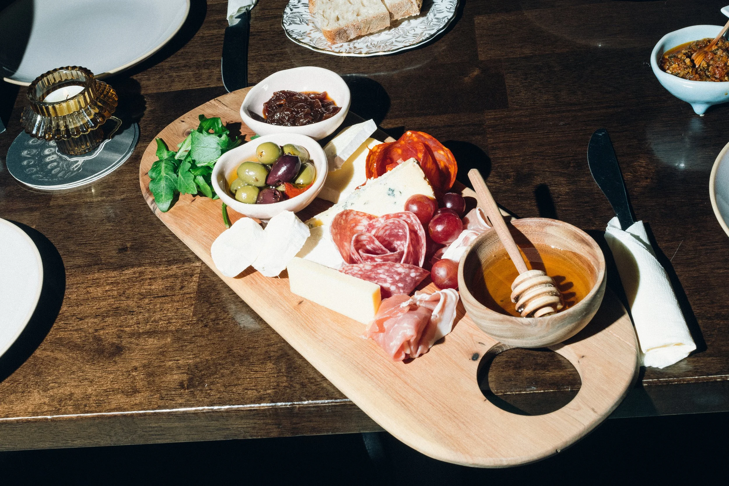 A cheese and charcuterie board on a wooden tray with various cheeses, cured meats, olives, honey, and small bowls of jams and spreads, on a dark wooden table.
