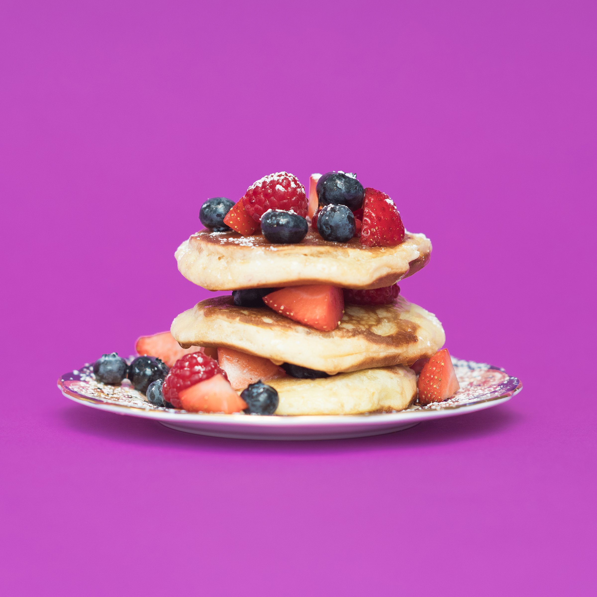 Stack of three pancakes topped with mixed berries including strawberries, blueberries, and raspberries, on a decorative plate against a purple background.