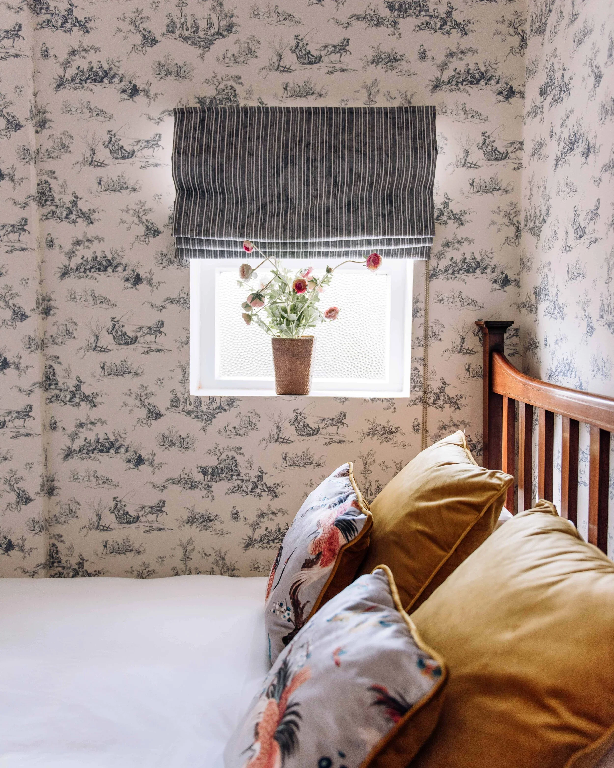A cozy bedroom with a bed, decorative pillows, a window with a striped valance, and a floral arrangement in a brown vase on the windowsill. The walls have toile de Jouy wallpaper with a pastoral scene.