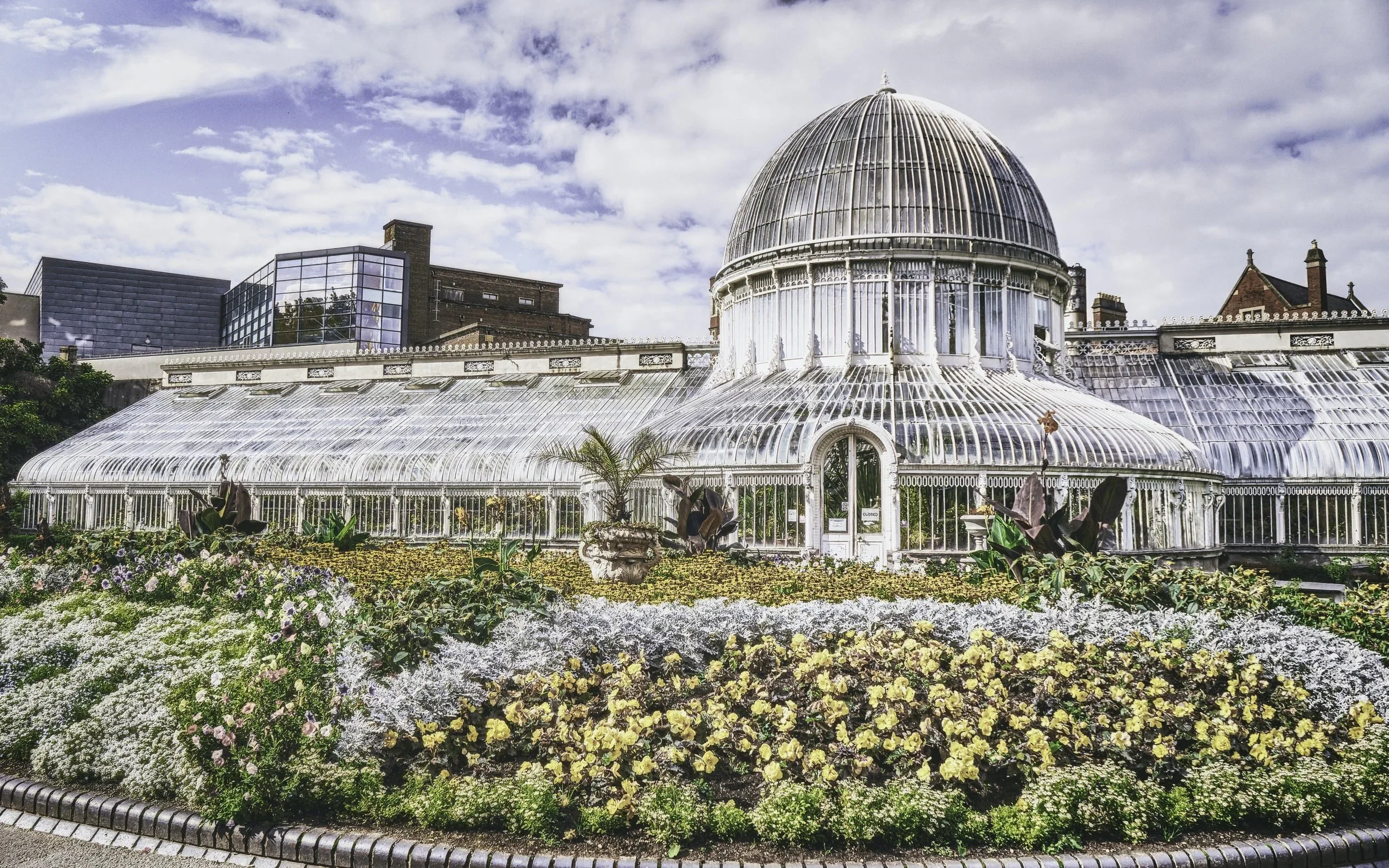 A large Victorian-style glass greenhouse with a dome roof, surrounded by colorful flower beds, under a partly cloudy sky.