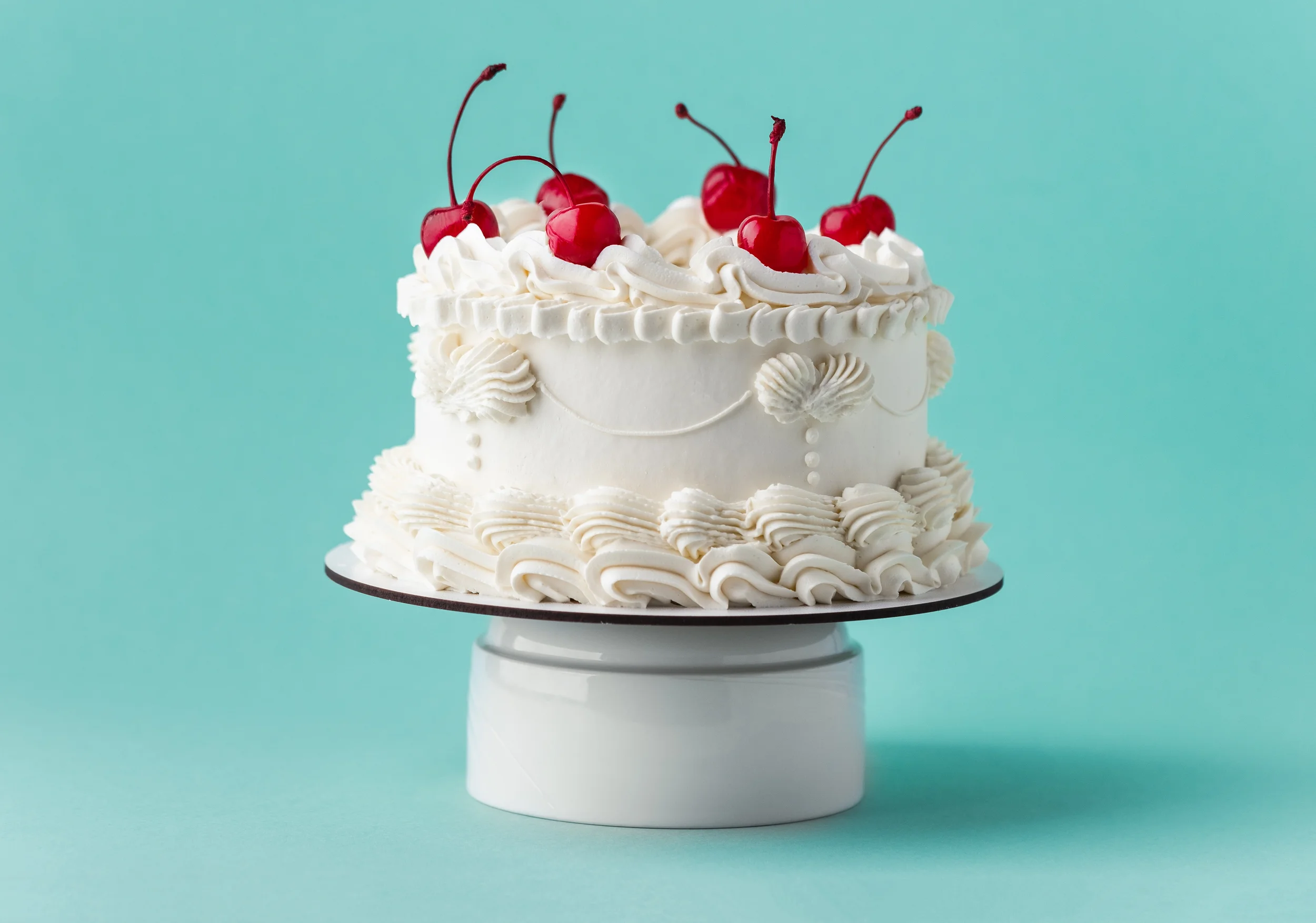 White frosted cake with cherries on top, decorated with whipped cream and piped icing, on a cake stand against a light blue background.