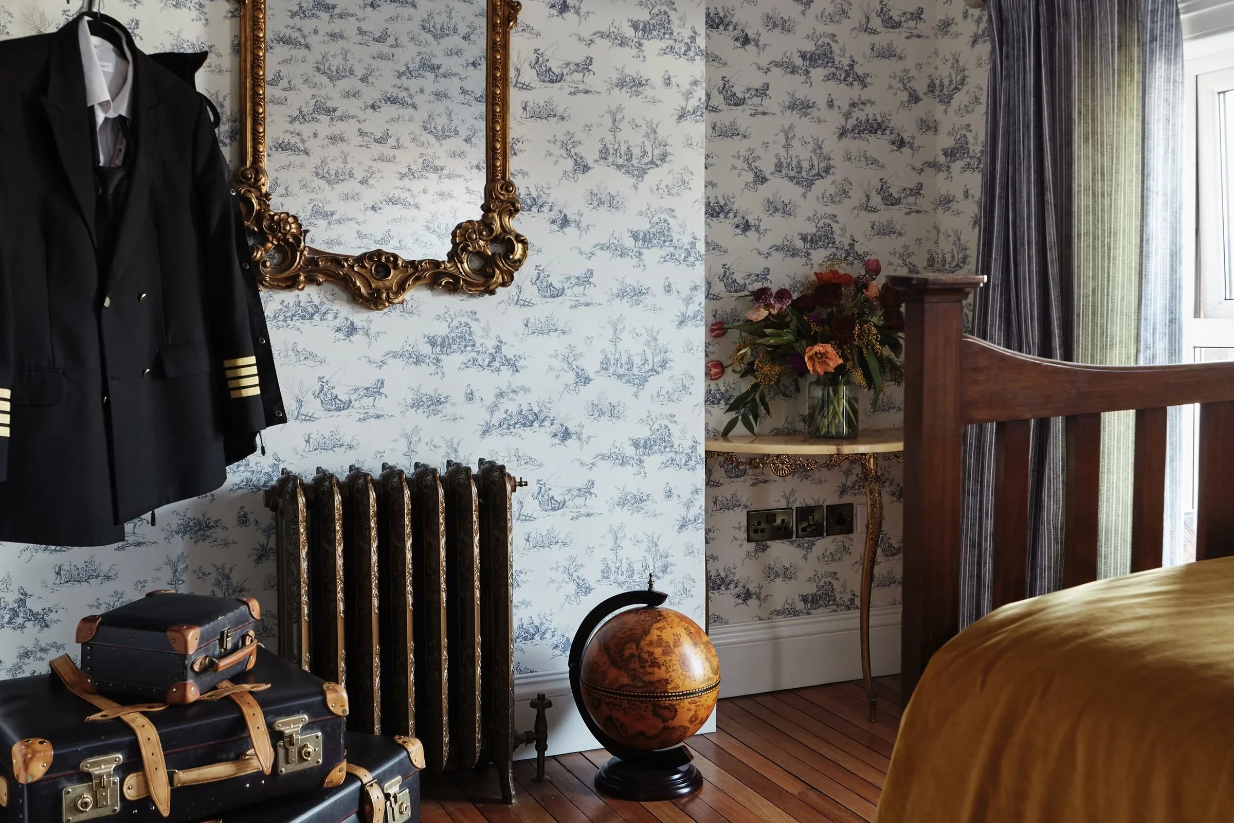 Interior of a bedroom with vintage decor, including a floral wallpapered wall, a gold ornate mirror, and a black radiator. There are suitcases on the floor, a globe next to the radiator, a side table with a vase of flowers, and a window with curtains