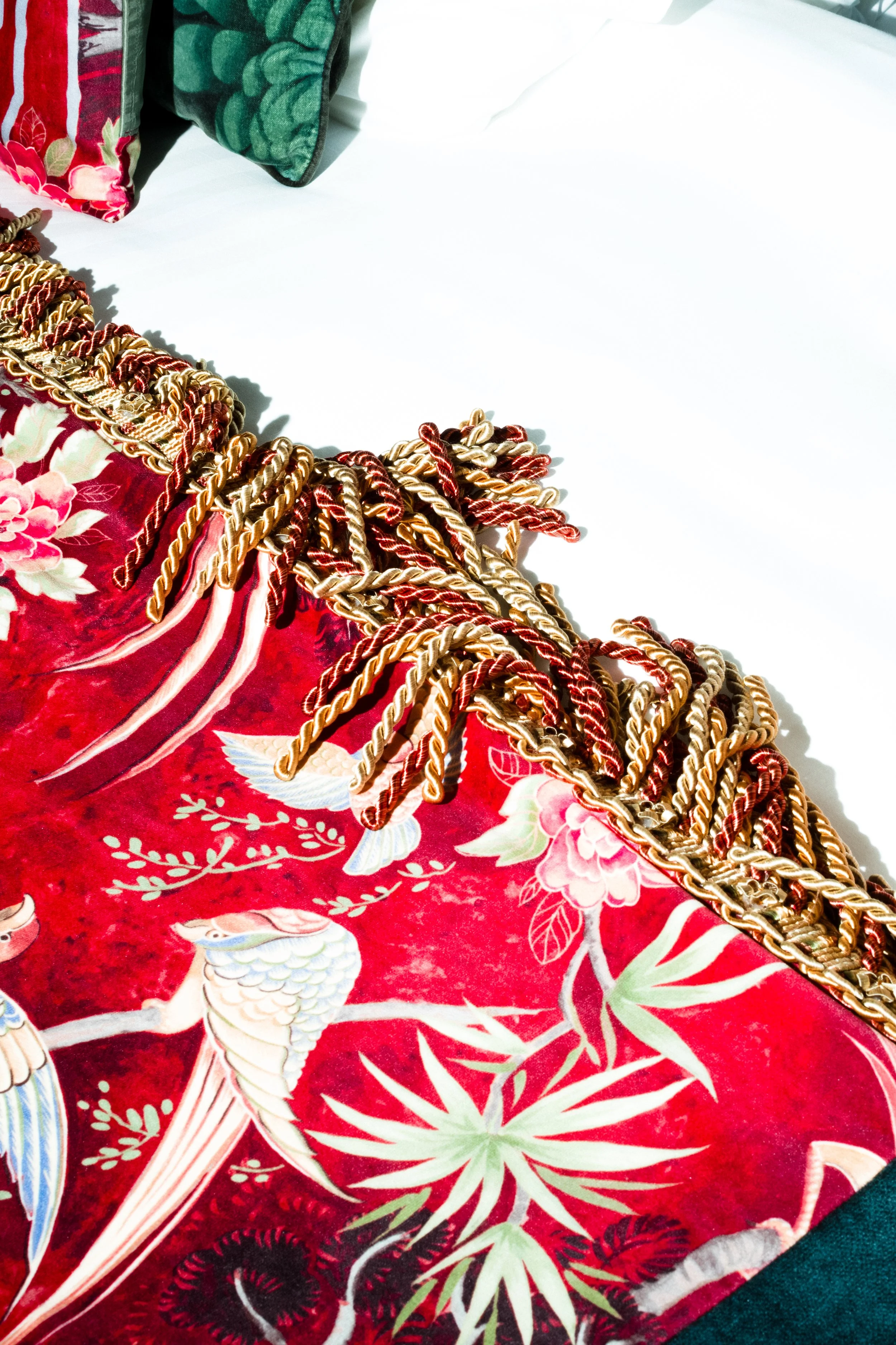 Close-up of a red fabric with floral and bird patterns, decorated with a multicolored braided fringe trim, placed on a white surface with pillows at the edge.