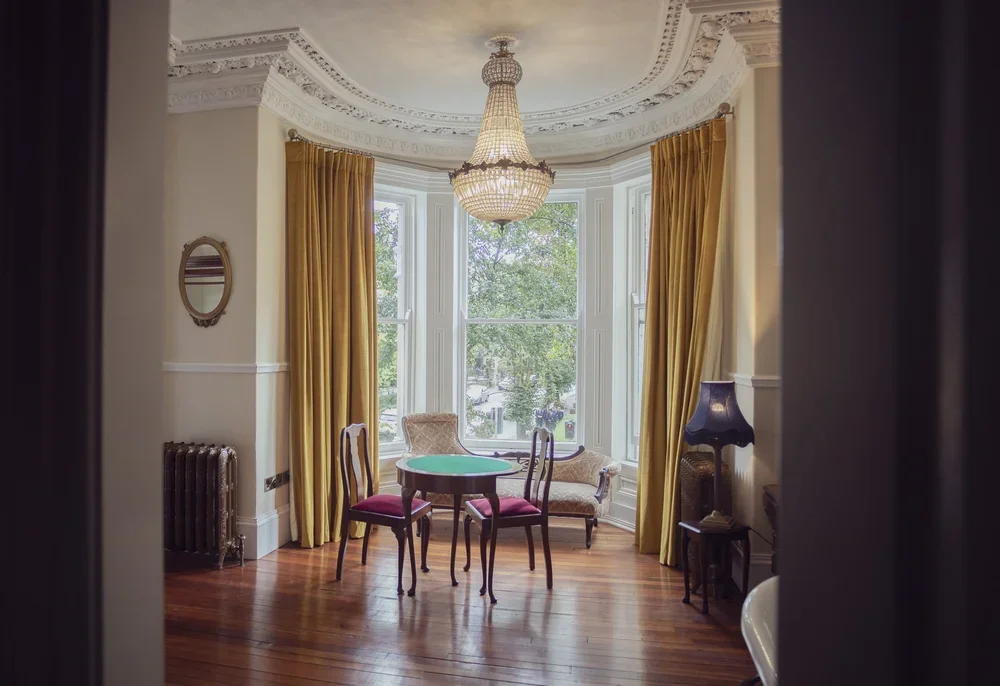 Elegant Victorian-style room with large bay window, gold curtains, a chandelier, wooden floor, small round table with four chairs, a vintage sofa, and a table lamp.