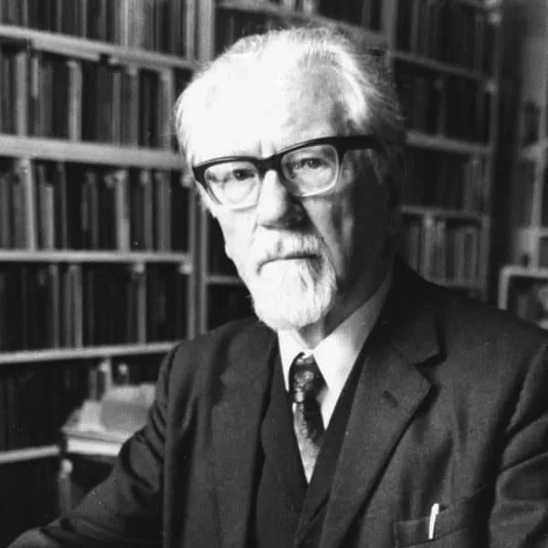 Black and white photo of an elderly man with white hair, beard, and glasses, wearing a suit and tie, sitting in front of a bookshelf filled with books.