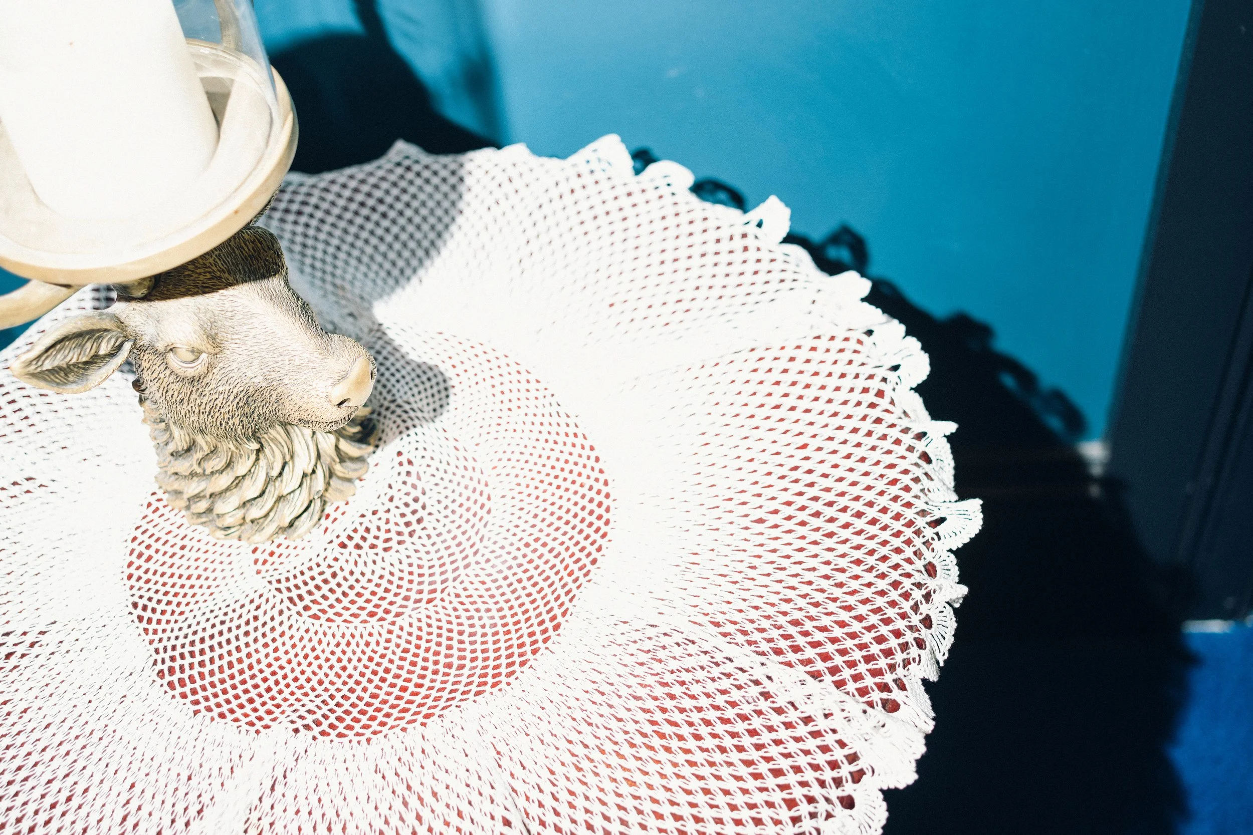 Close-up of a decorative table with a white lace runner, a gnome figure with a sheep's head, and a white candle in a glass container. The background features a blue wall.