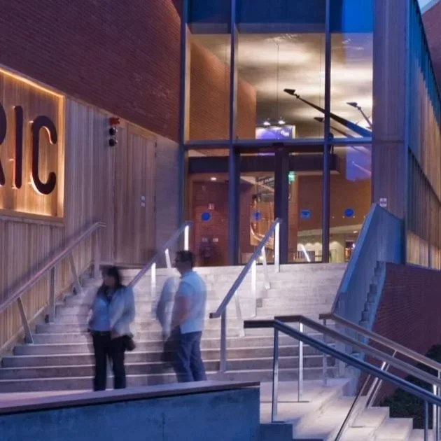Two people standing on the stairs outside a modern building with glass walls, illuminated sign, and wooden paneling, possibly entering or exiting.