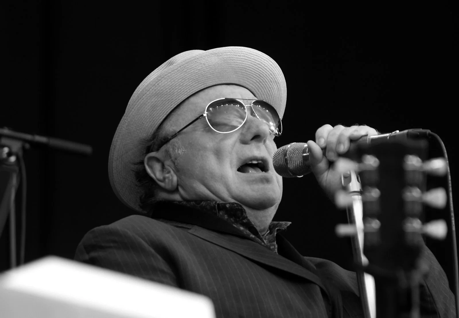 A black and white photo of a man singing into a microphone, wearing sunglasses, a wide-brimmed hat, and a suit.