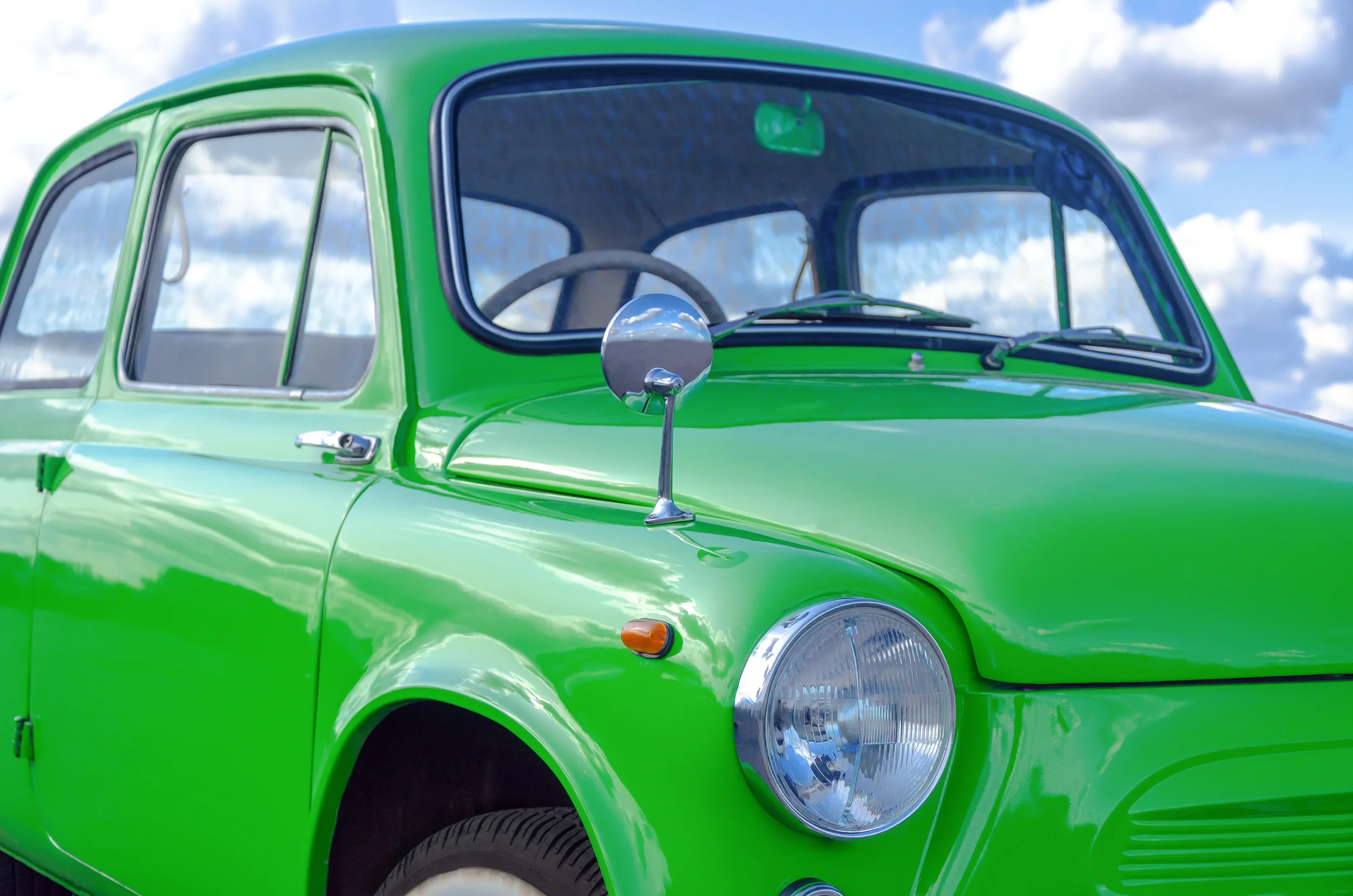 A bright green vintage compact car with rounded edges, a large headlight, side mirror, and windows reflecting a cloudy blue sky.