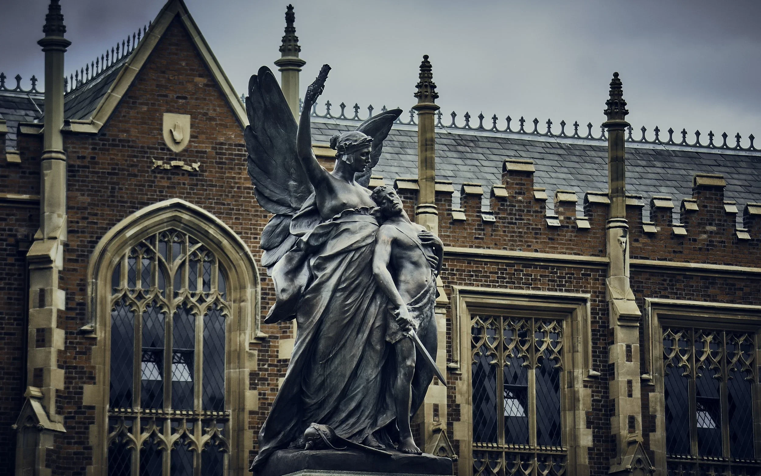 A bronze statue of a woman with wings, holding a sword and standing over a man, in front of a historic brick building with large arched windows.