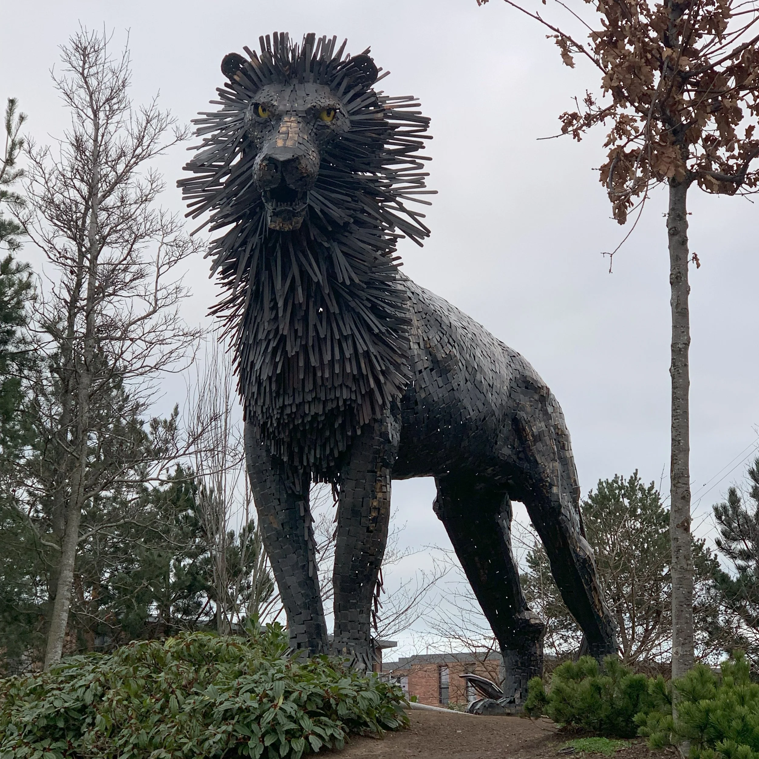 Large black lion statue made of metal standing outdoors among trees and bushes.