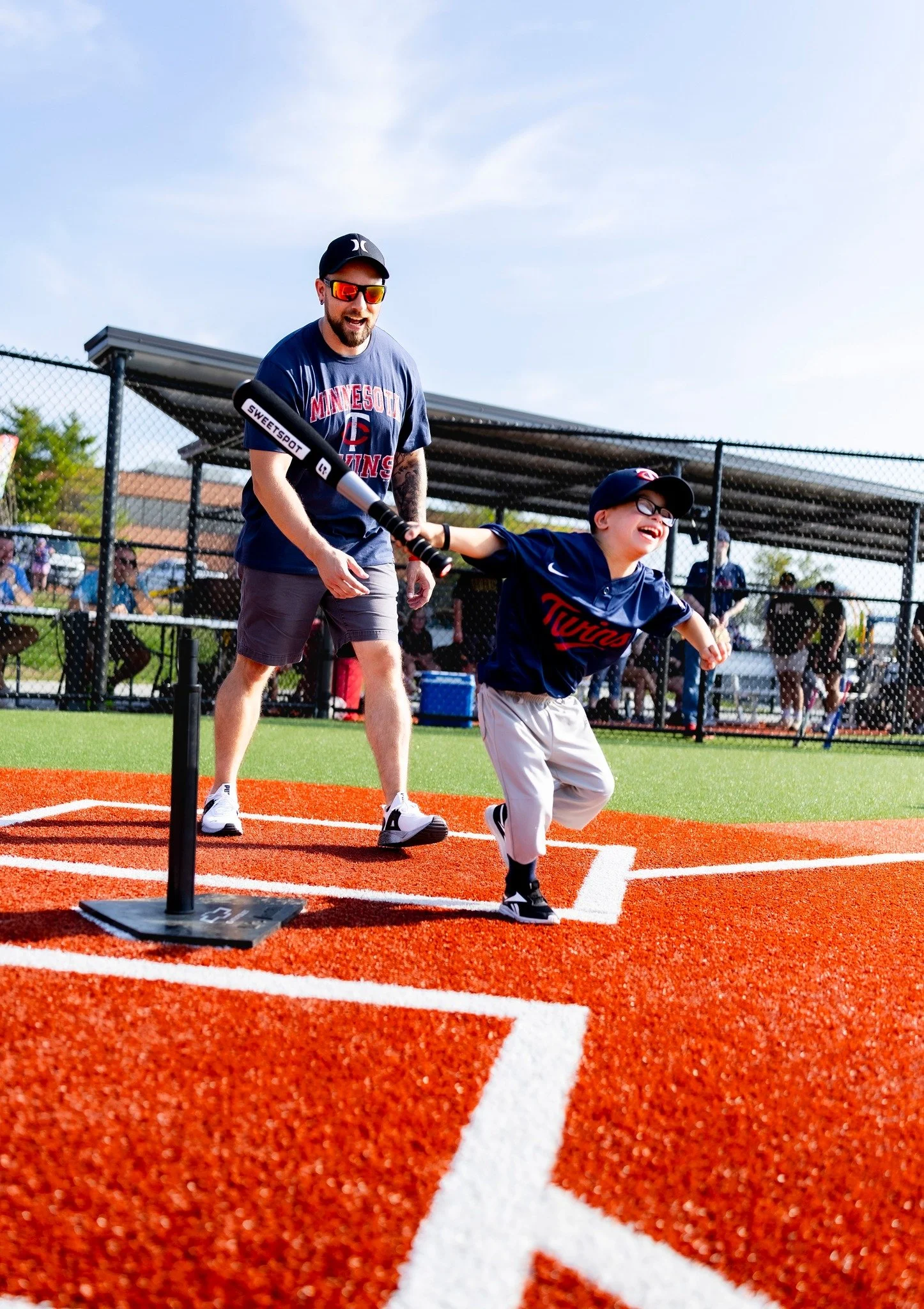A young boy in a baseball uniform running home as an adult man in casual clothing hits a baseball with a bat during a youth baseball game on a sunny day.