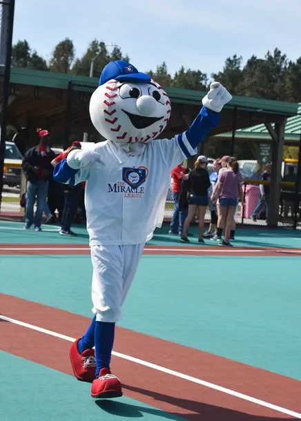 Mascot dressed as a baseball with a face, wearing a blue cap, white uniform with 'Miracle League' logo, red shoes, and blue and white socks, walking on a baseball field with people in the background.