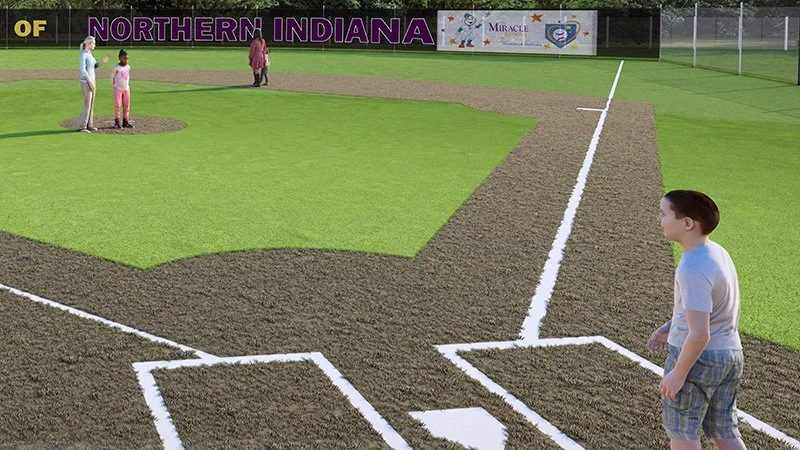 Child standing on a baseball field infield near the home plate, watching others near the outfield. The field has green grass and brown dirt with white chalk lines, with a sign reading 'Northern Indiana' in the background.