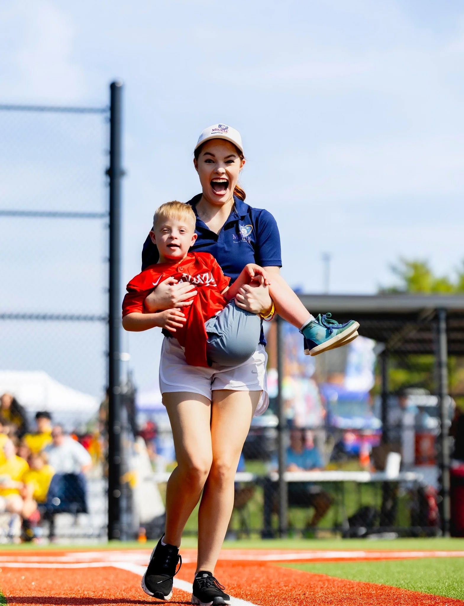 A woman wearing a navy blue uniform and a white cap holding a young boy in a red sports jersey on a sports field, both appearing happy and excited.
