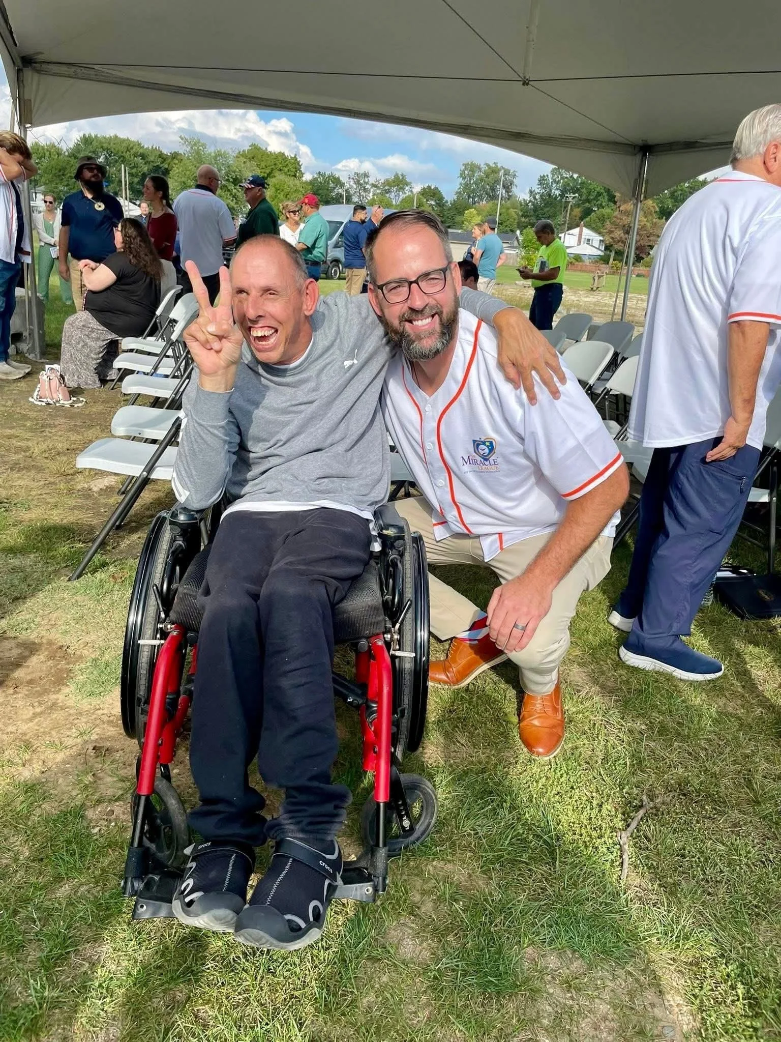 Two men smiling and hugging, one in a wheelchair, at an outdoor event under a canopy with other people in the background.