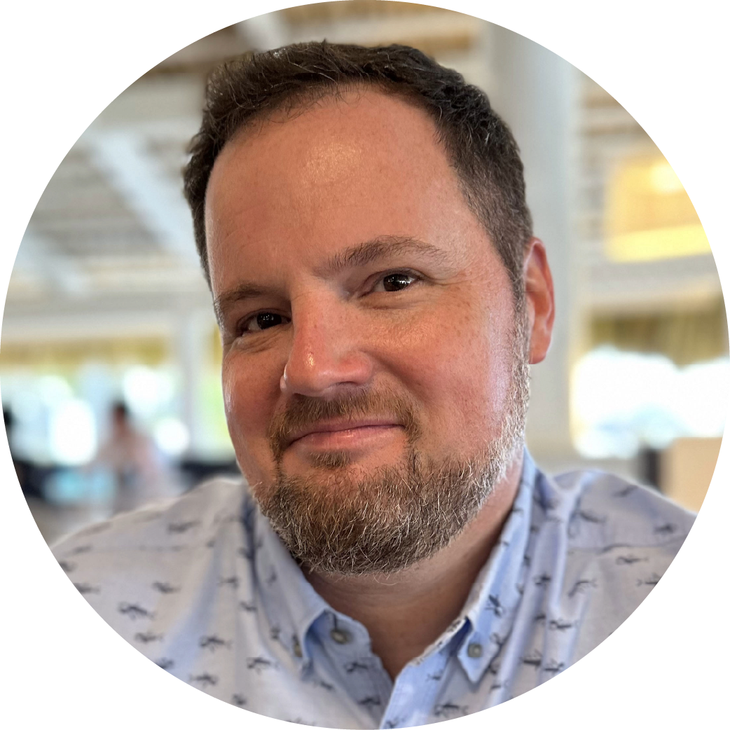Close-up of a man with short dark hair, a beard, and a mustache, smiling slightly, wearing a light-colored patterned shirt, in a bright indoor setting with blurred background.