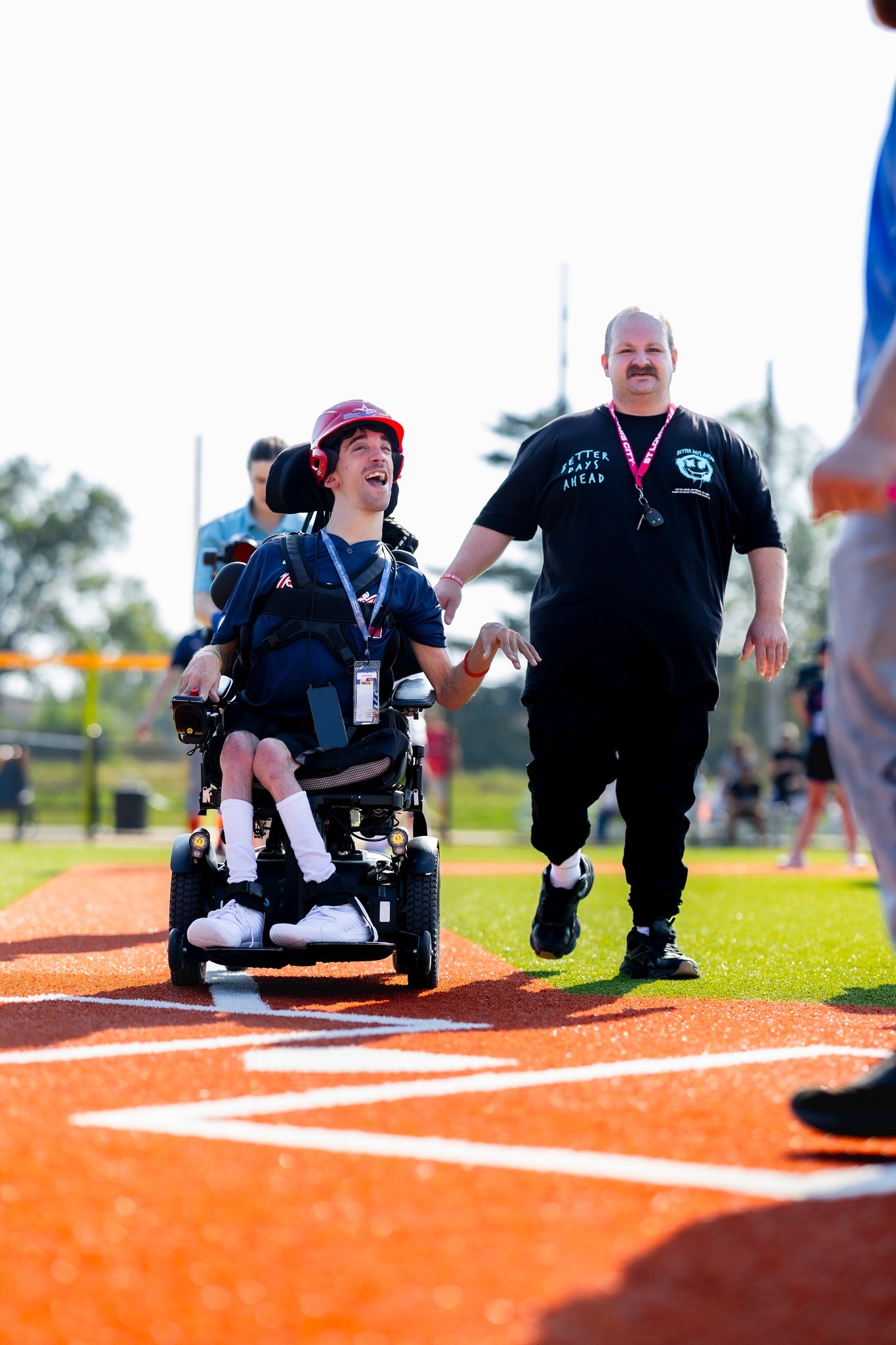 A young man in a wheelchair smiling and holding hands with a man running on a sports track during an outdoor event on a sunny day.