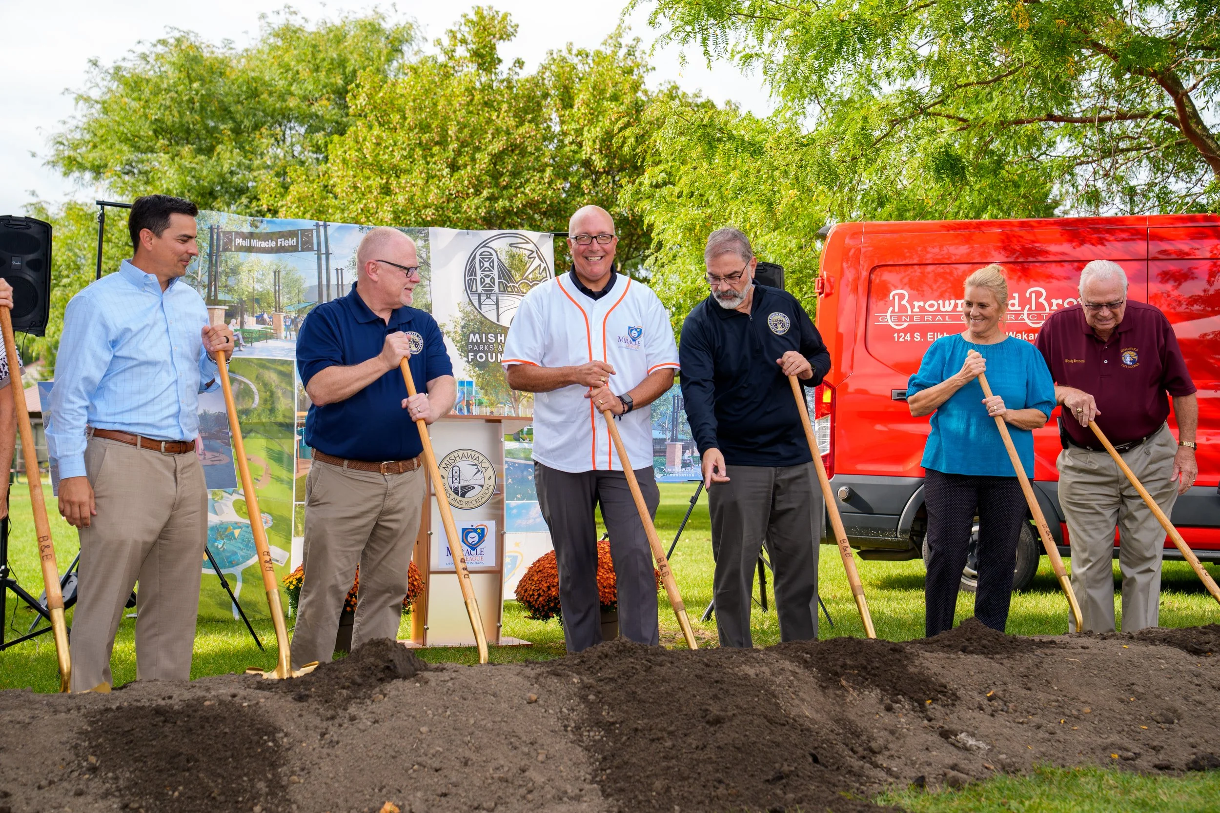 A group of seven people, some holding shovels, participating in a groundbreaking ceremony outdoors. They are standing behind a mound of dirt with a backdrop showing park development plans, surrounded by green trees and a bright sky.