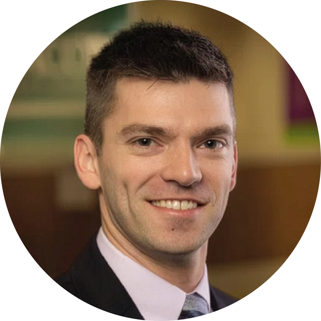 A professional man with short dark hair, wearing a dark suit and tie, smiling in an indoor setting with blurred background.