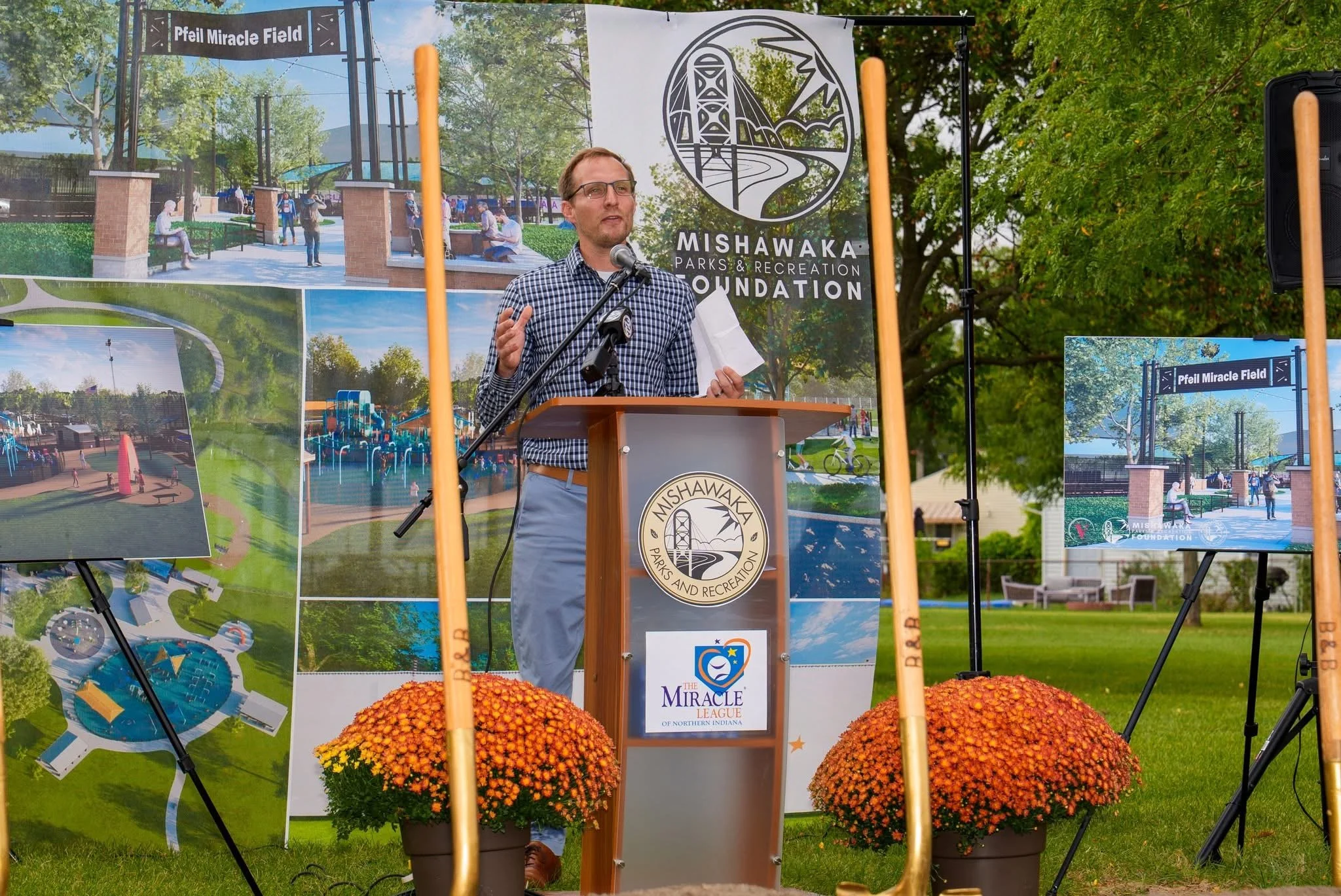 A man wearing glasses, a checkered shirt, and gray pants speaking at a podium outdoors during a public event. Behind him are banners and posters showing a park design and the logo of Mishawaka Parks & Recreation Foundation. There are orange potted flowers in front of the podium and trees in the background.