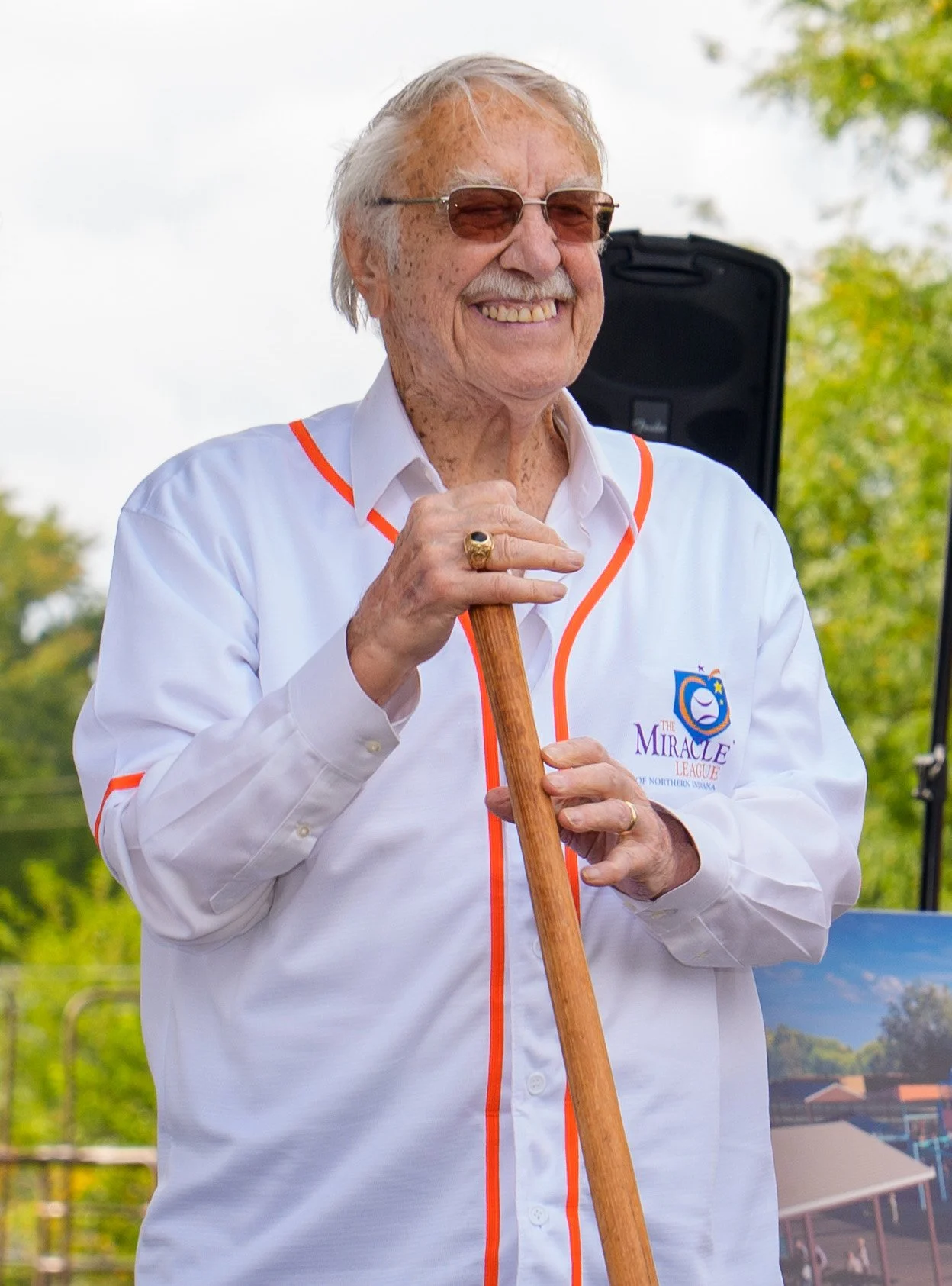 An elderly man smiling outdoors, wearing sunglasses and a white shirt with orange piping, holding a wooden cane, with trees and a speaker in the background.