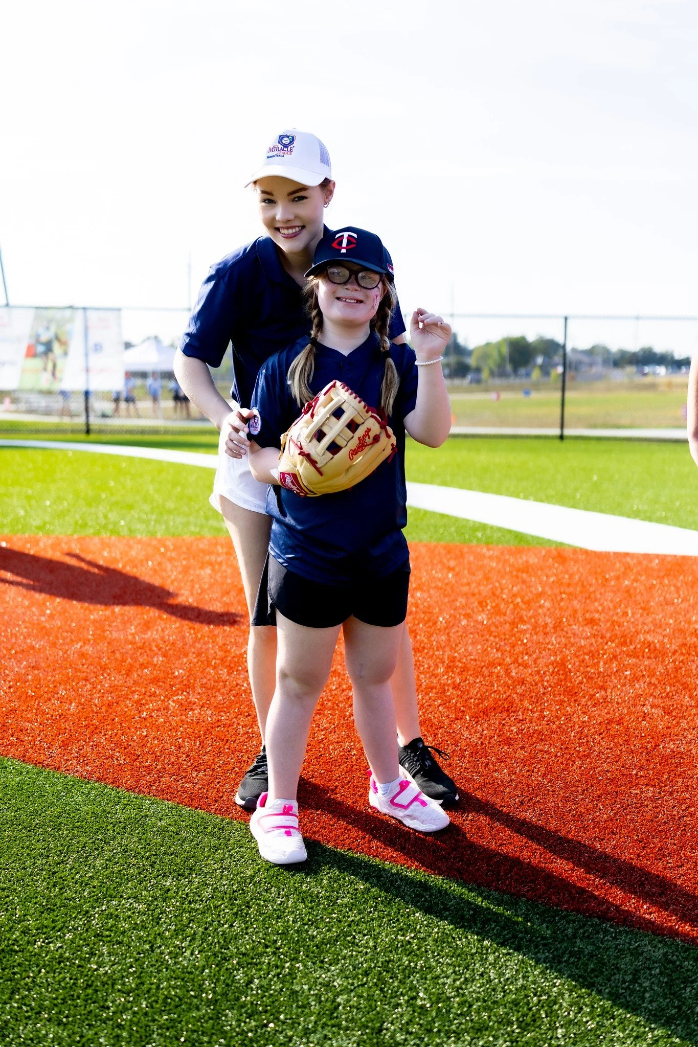 A young girl and a woman are standing on a baseball field, smiling at the camera. The girl is wearing glasses, a navy baseball cap, a navy shirt, shorts, and sneakers, holding a baseball glove. The woman behind her is also smiling, wearing a white cap and navy shirt, standing on the grass and red clay of the field.