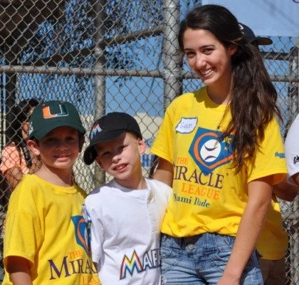 Three people, two children and one young woman, standing outdoors in front of a chain-link fence. The children are wearing yellow T-shirts, and the young woman is also dressed casually, smiling at the camera.