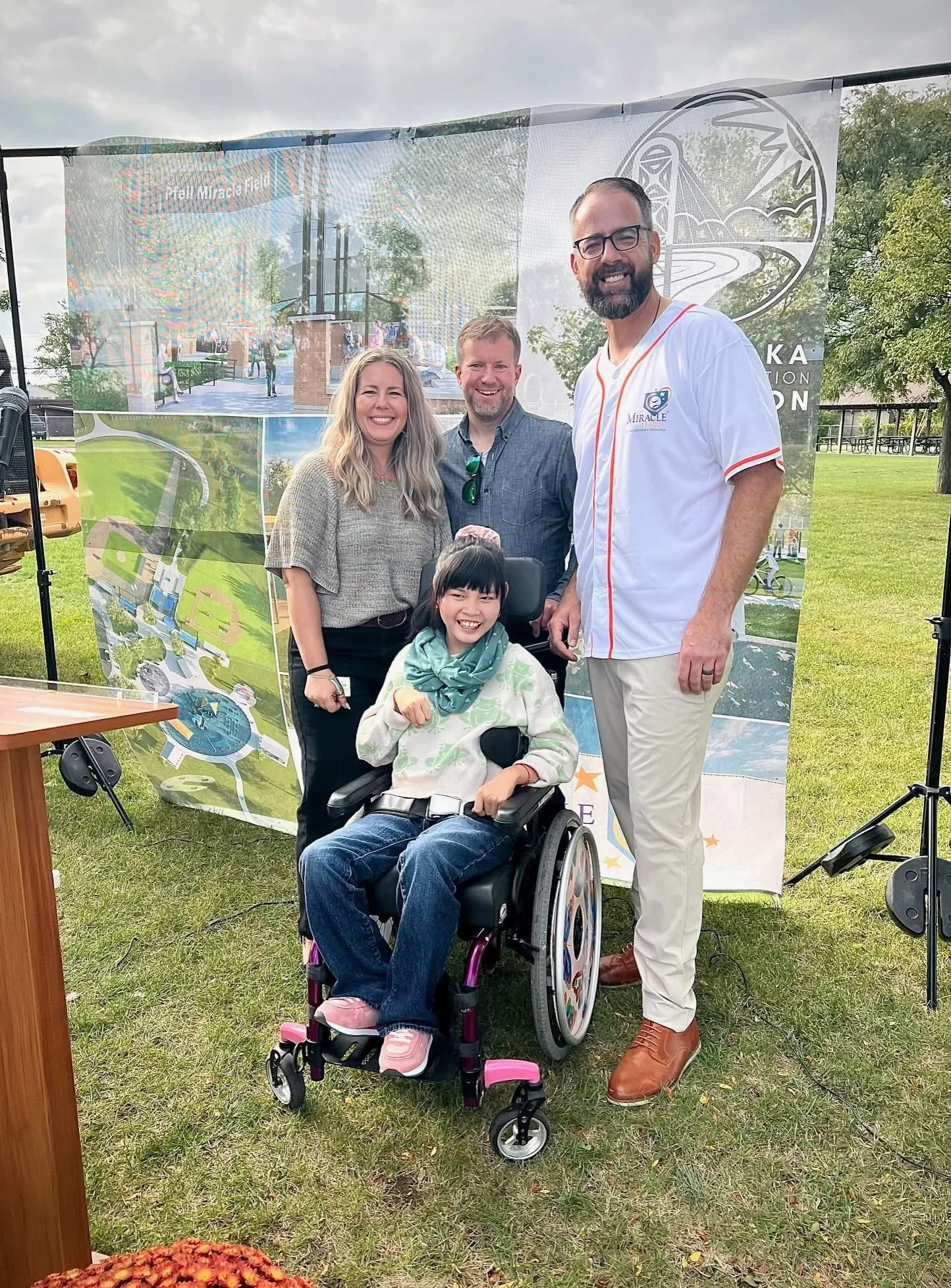 A group of four adults and one young girl in a wheelchair posing outdoors. The girl is smiling, wearing a cream hoodie with green patterns, and pink shoes. The background features a colorful banner, trees, and a grassy park.