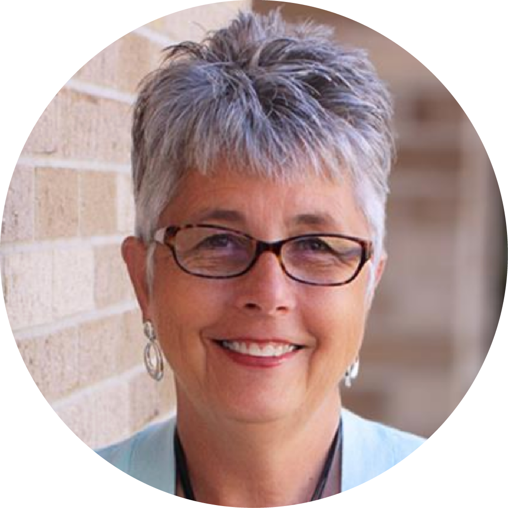 Portrait of a smiling middle-aged woman with short gray hair and glasses, standing against a brick wall.
