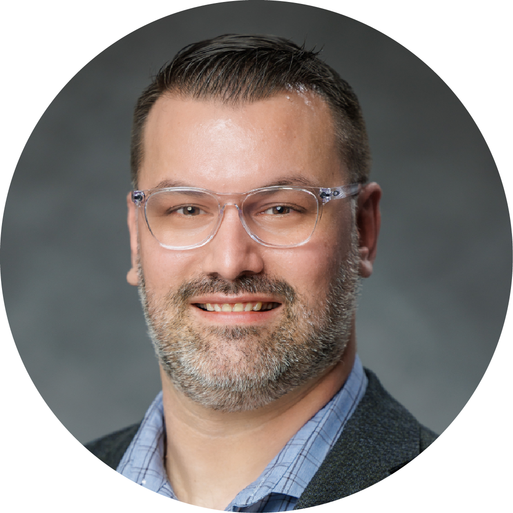 A professional headshot of a smiling man with glasses, short dark hair, and a beard, wearing a blue shirt and a dark blazer against a gray background.