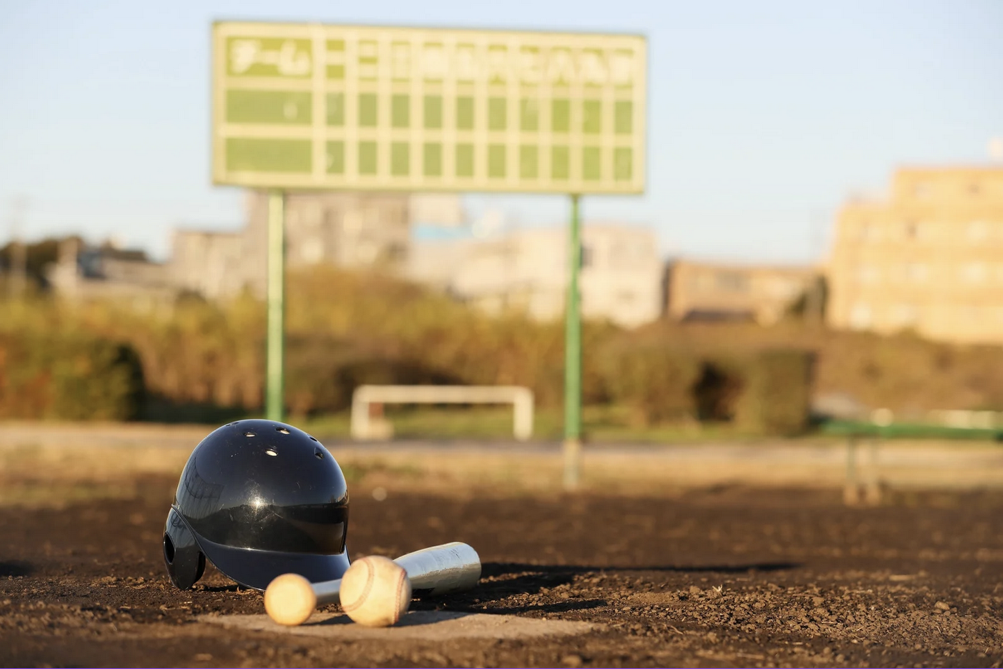 A baseball helmet, a bat, and two baseballs on the ground of a baseball field, with a blurry soccer goal and scoreboard in the background during daylight.