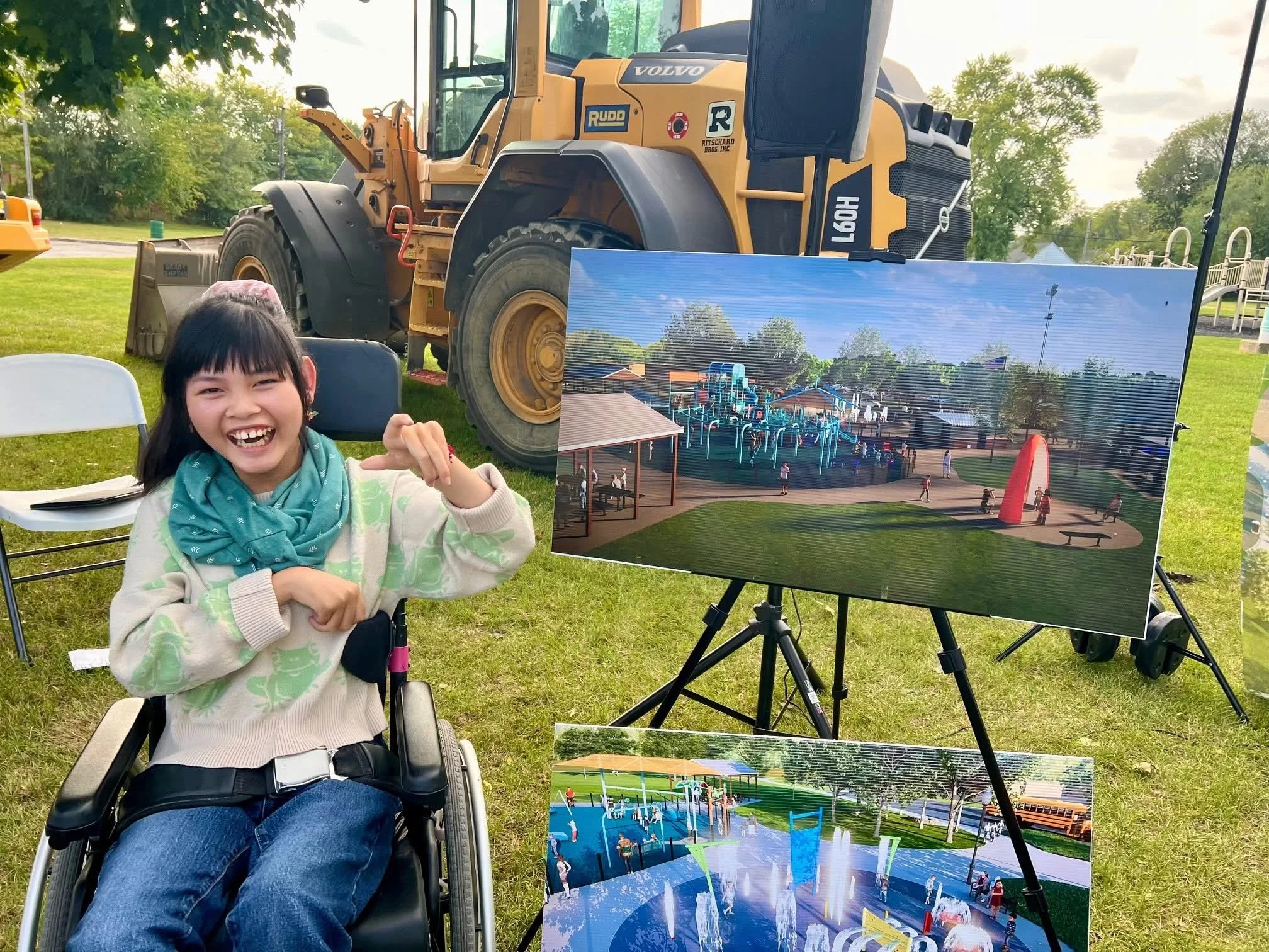 A young girl with black hair, wearing a beige sweater with green patterns, a teal scarf, and blue jeans, seated in a wheelchair, smiling and pointing at an outdoor art display. Behind her, there are two large prints of colorful park scenes on easels, a large yellow Volvo construction vehicle, and a green park with trees.