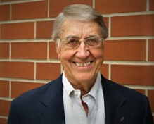 A smiling elderly man with gray hair and glasses, standing in front of a brick wall, dressed in a suit and tie.