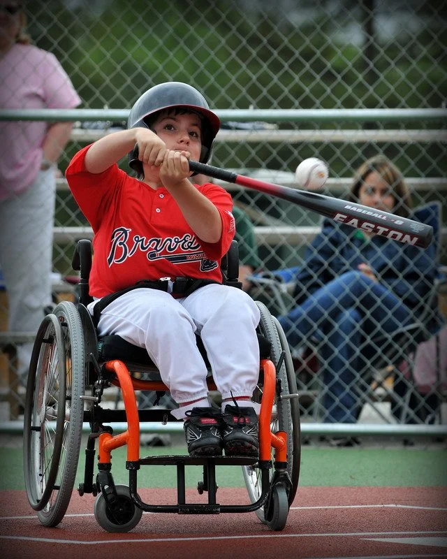 Young boy in a wheelchair wearing a baseball cap, red jersey, and white pants, swinging a baseball bat at a ball on a baseball field.