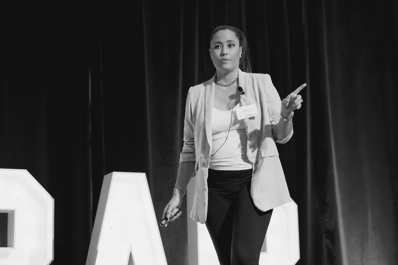 A woman speaking on stage during a presentation, with large illuminated letters in the background.