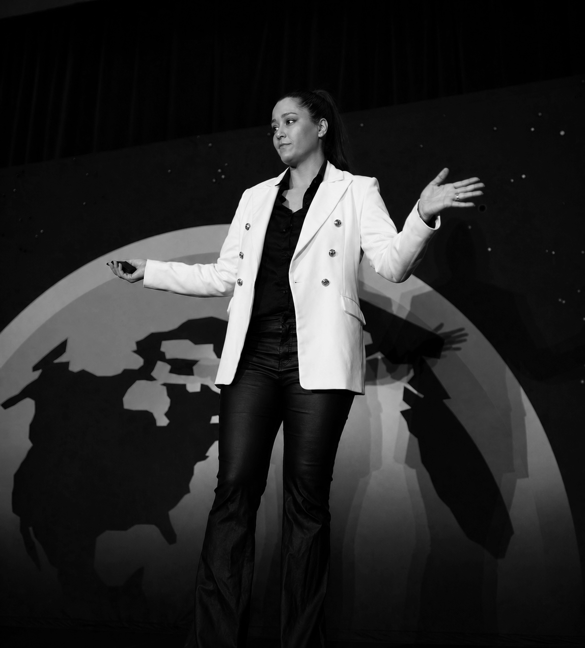 A woman giving a presentation on stage with a large globe background, wearing a white blazer and black pants.